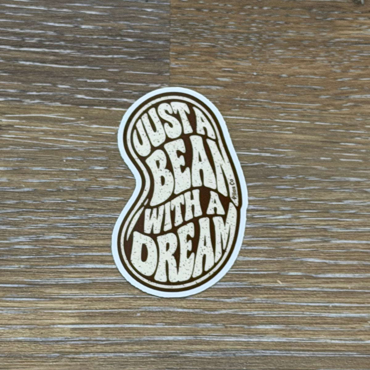 A die-cut sticker shaped like a bean with the text 'JUST A BEAN WITH A DREAM' in a retro bubbly font, placed on a brown wood-grain surface.