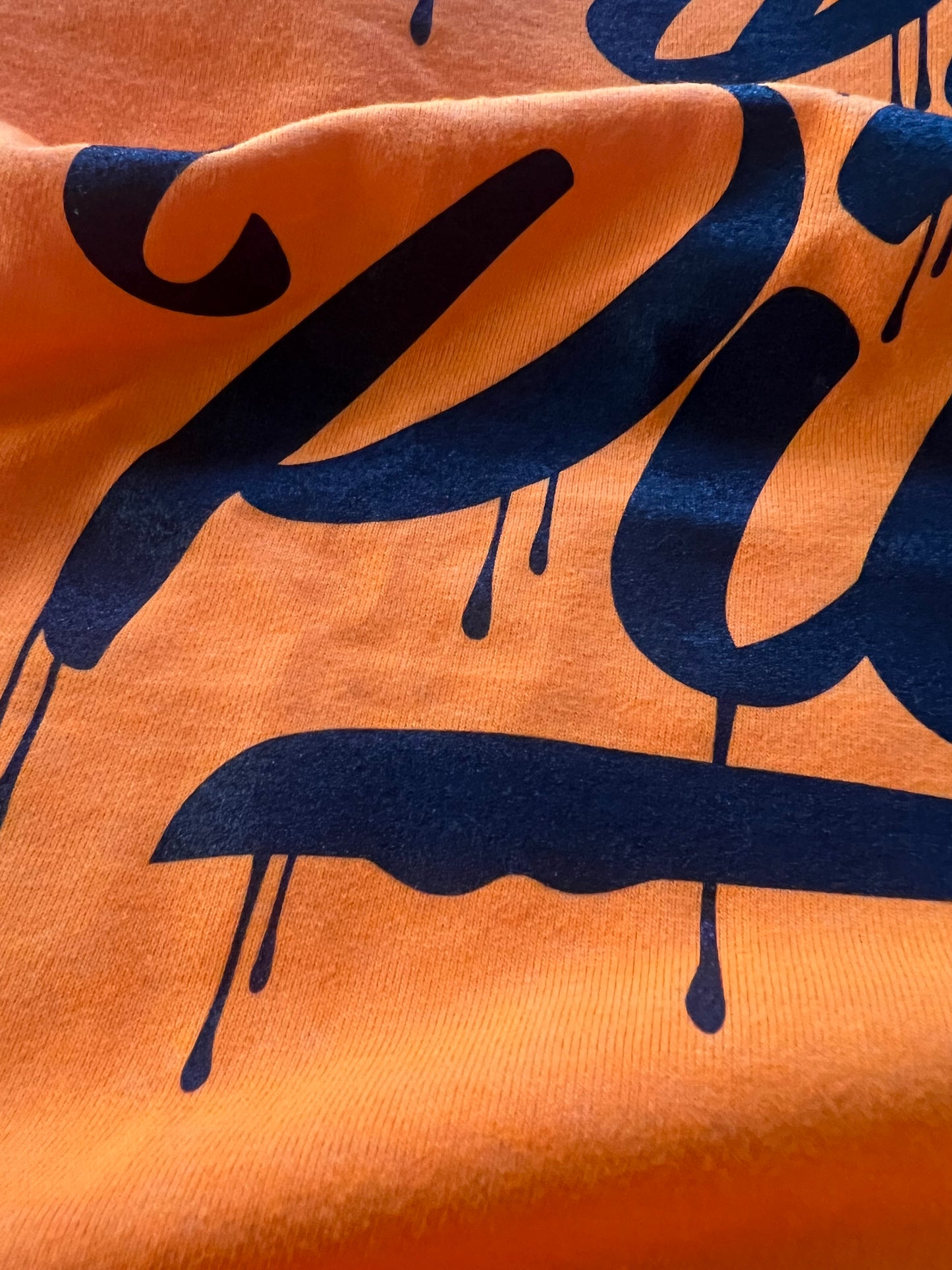Close-up detail of Pilas Clothing Co. PLP Drip Hi-Vis safety shirt in bright orange with black graffiti-style drip lettering, showing fabric texture and print quality