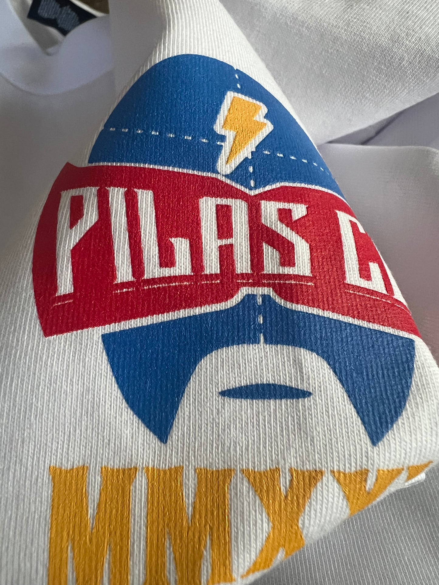 Close-up detail of Pilas Clothing Co. logo shirt in white featuring blue luchador mask with lightning bolt, red banner with PILAS text, and gold/yellow accents on premium cotton fabric showcasing vibrant screen print quality