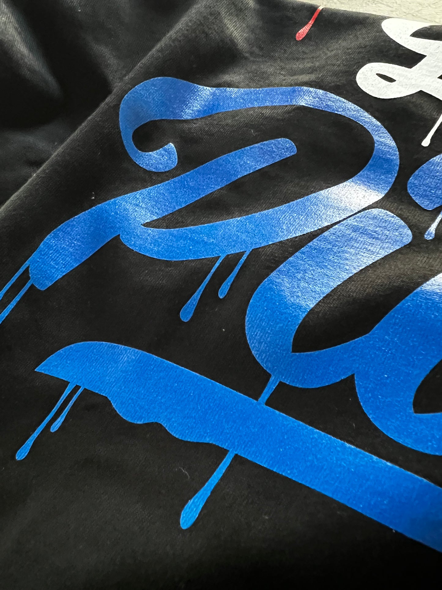 Close-up detail of Pilas Clothing Co. PLP Drip USA Flag shirt in black with bold blue graffiti-style drip lettering, showcasing premium cotton fabric and vibrant screen print quality inspired by American flag colors