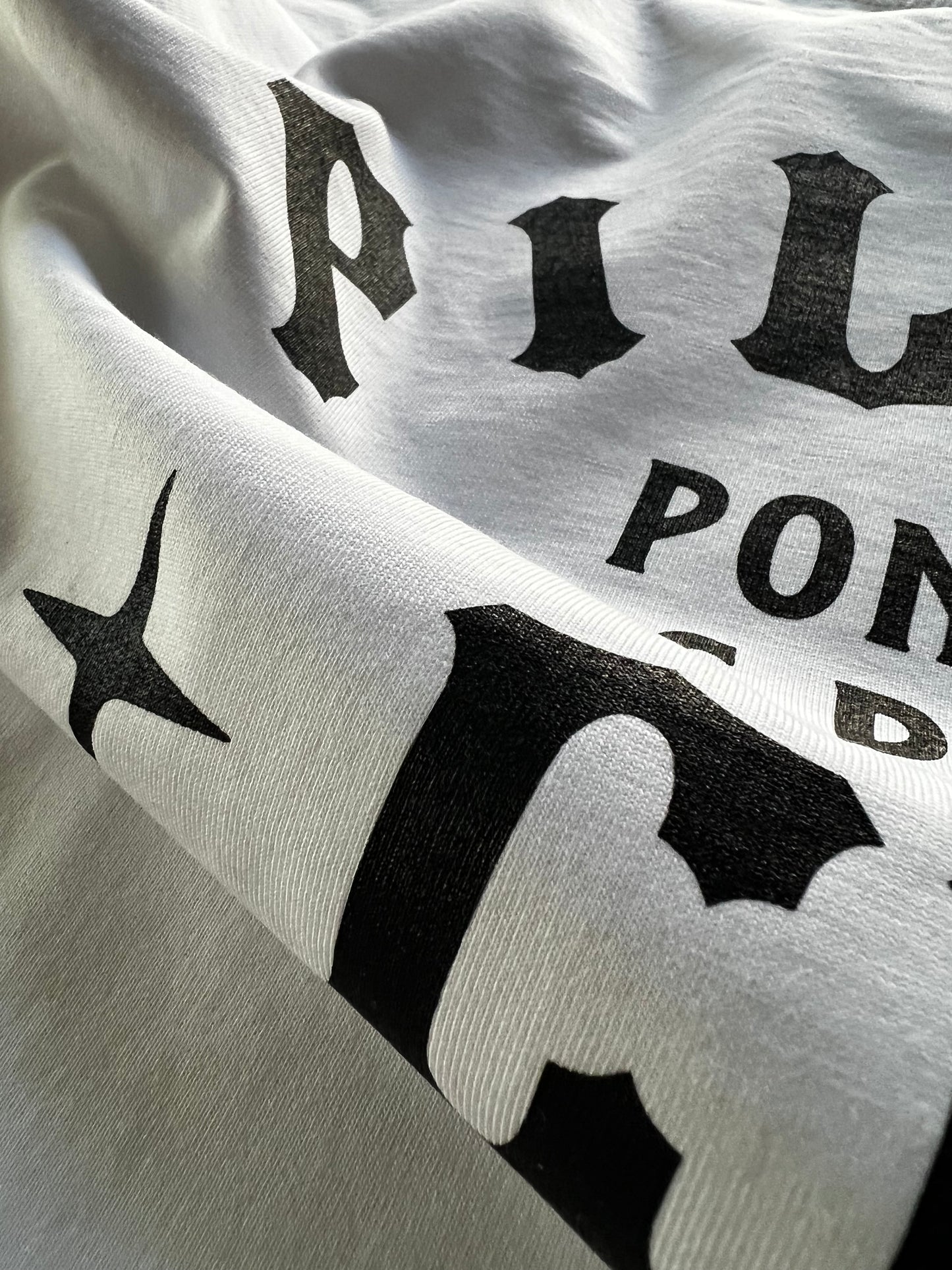 Close-up detail of Pilas Clothing Co. PLP Ponte Las Pilas text shirt in white with black gothic-style lettering, showcasing premium cotton fabric texture and crisp screen print quality