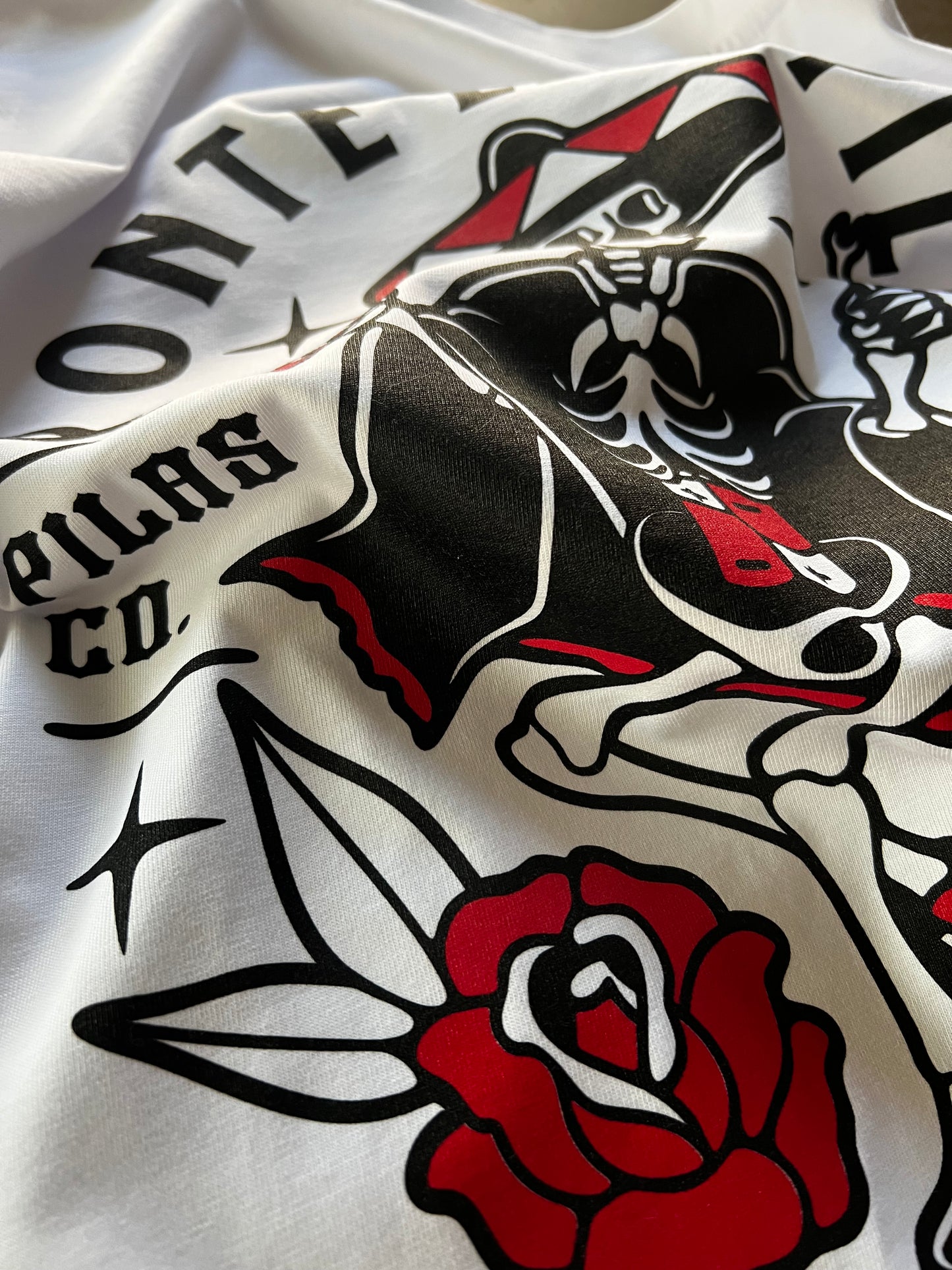 Detail shot of Pilas Clothing Co. Mueve El Esqueleto dancing skeleton shirt in white with bold black and red tattoo-inspired design showcasing Day of the Dead skull, roses, and traditional artwork