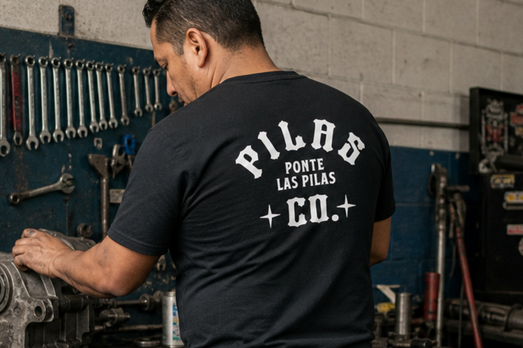 A man stands with his back to the camera in a garage workshop, wearing a black t-shirt with 'PILAS PORTE LAS PILAS CO.' printed in white on the back.