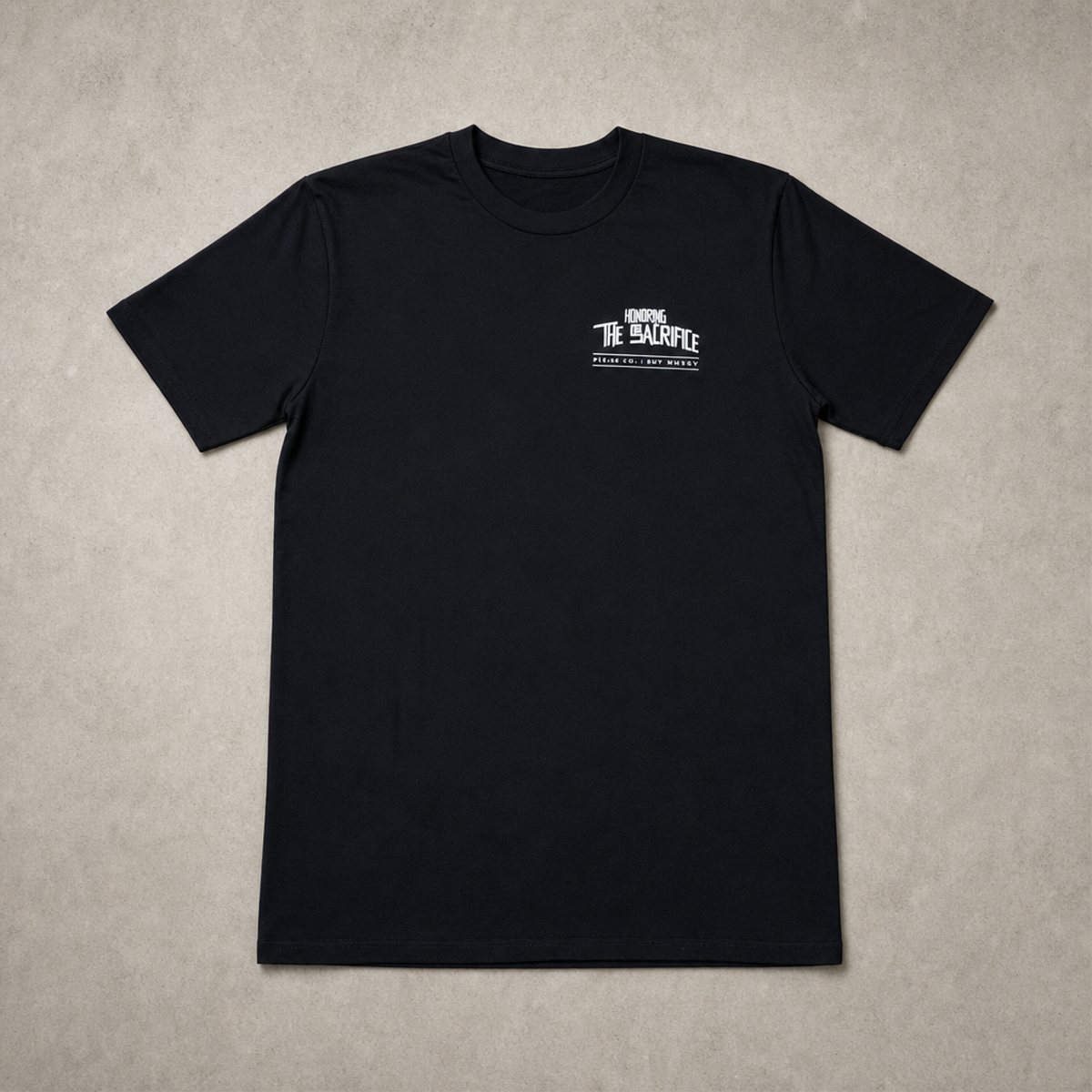 A black short-sleeve crewneck t-shirt with a small white graphic on the left chest reading 'honoring the sacrifice' and 'Pilas co' 'EST. 2024', laid flat on a textured gray background.