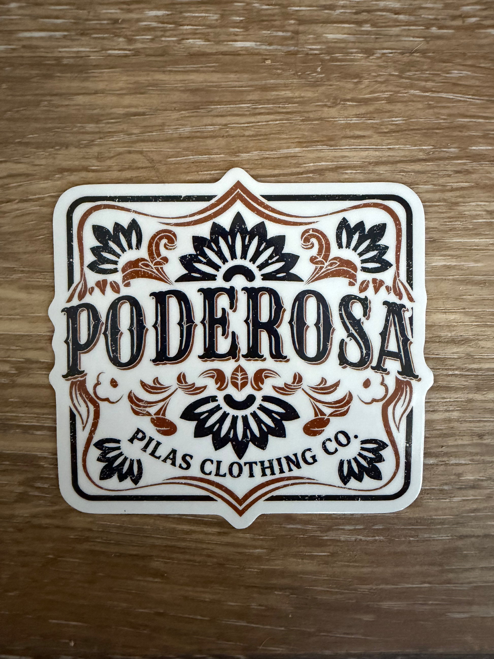 Single 3.5 inch Poderosa die-cut vinyl sticker with black and copper western lettering and Talavera floral design, laid on a wood surface, by Pilas Clothing Co.