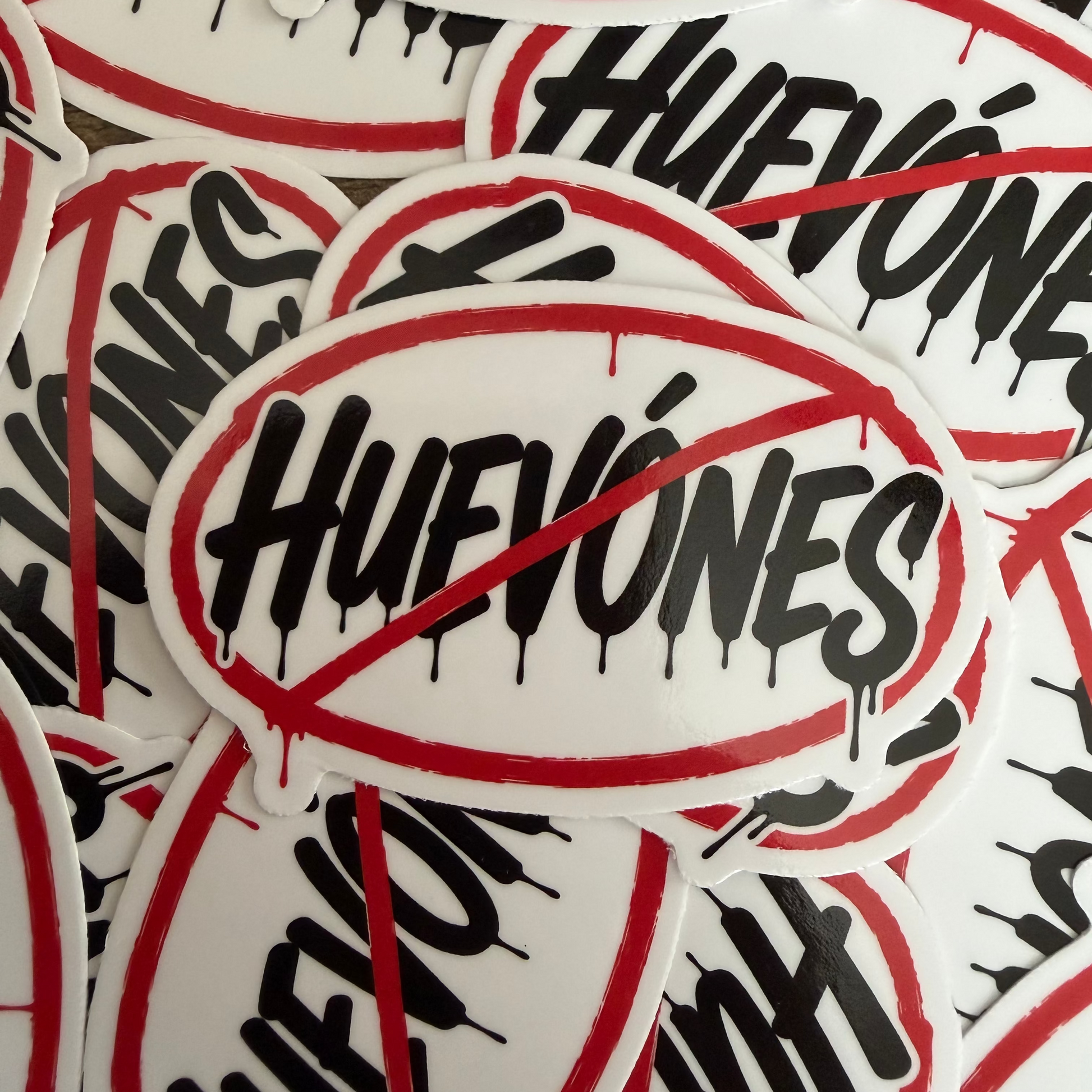 A pile of white oval stickers with the word 'HUEVONES' in black script crossed out with a red diagonal prohibition symbol.
