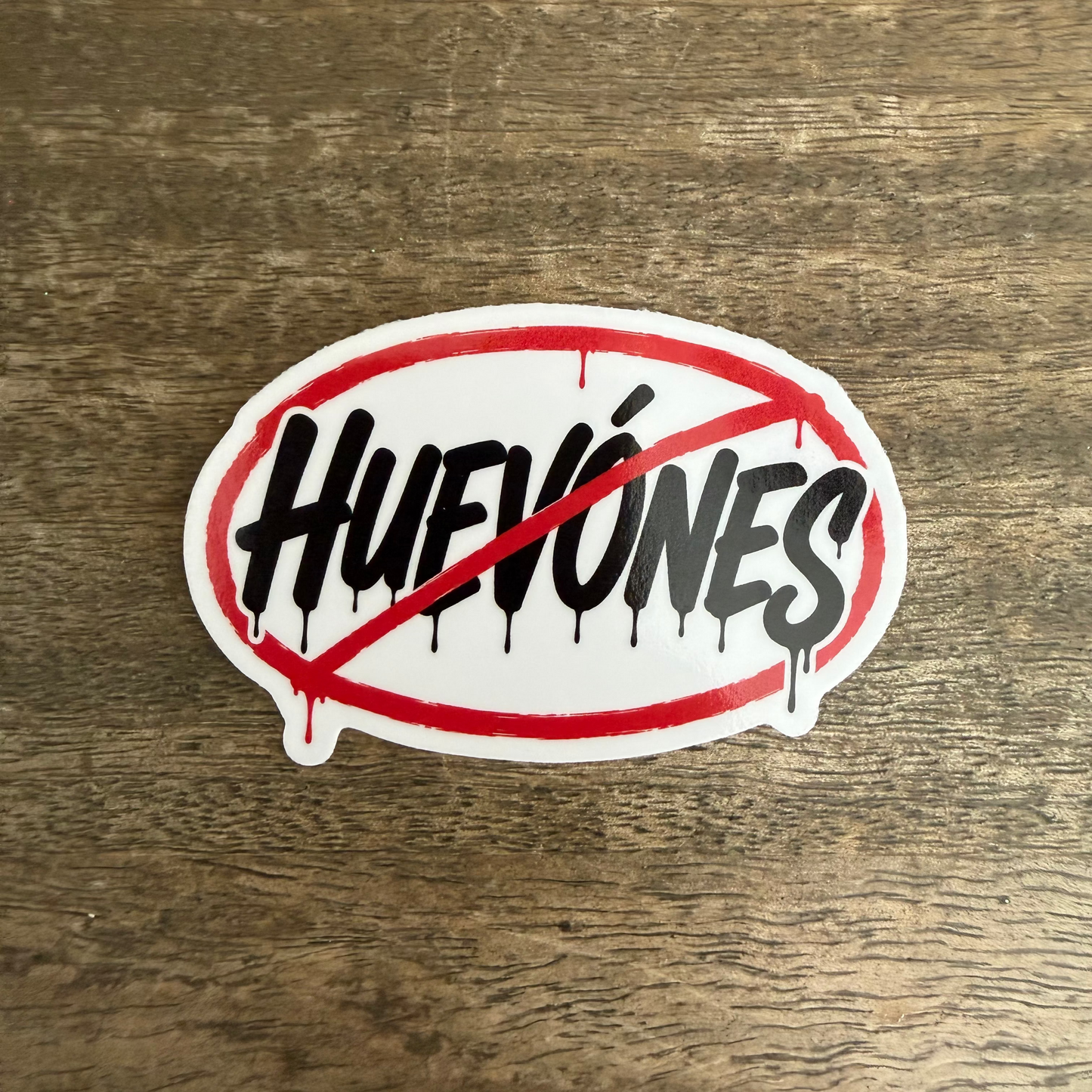 An oval sticker with the word 'HUEVONES' in black dripping-style lettering, crossed out by a red diagonal line and red oval border, placed on a brown wood surface.