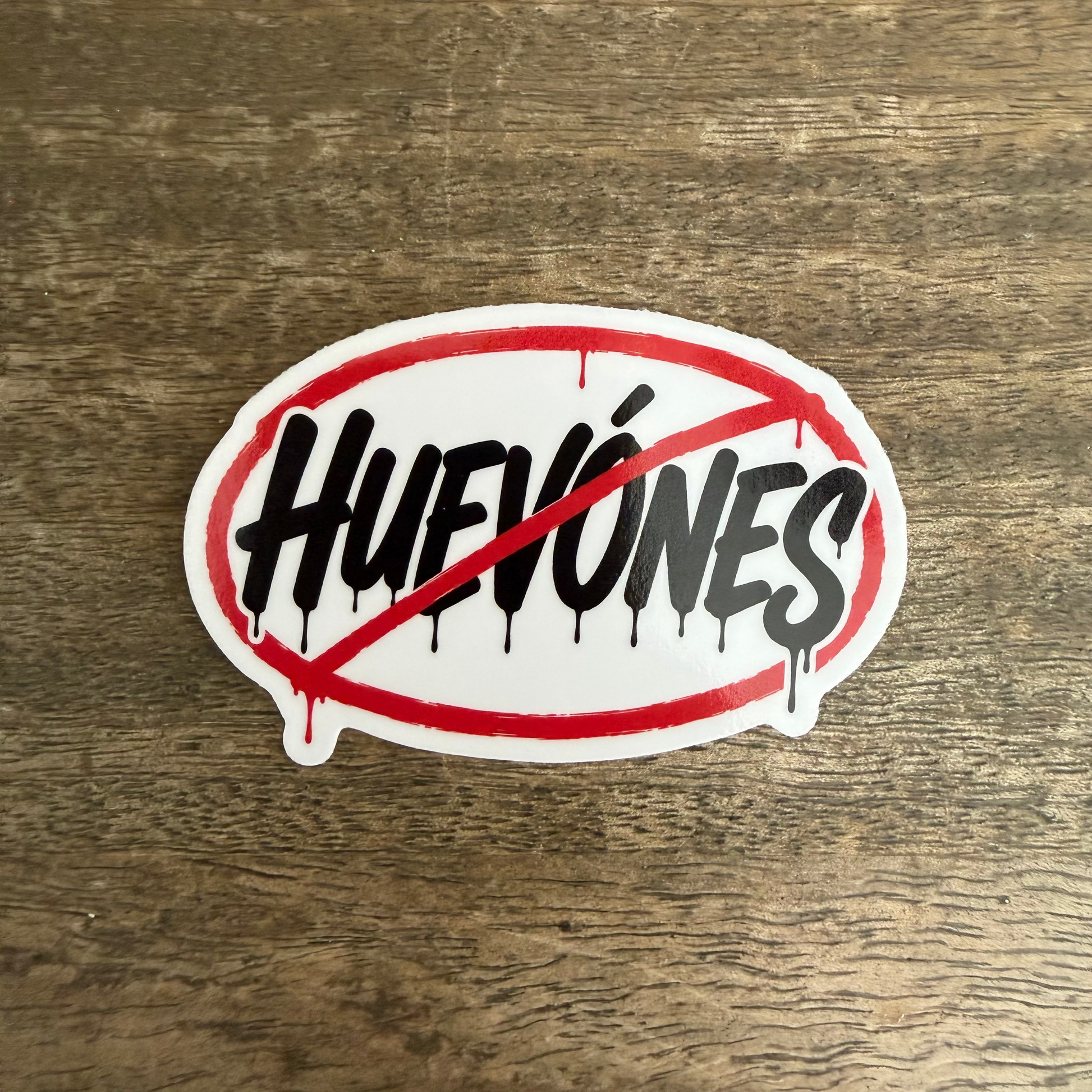 An oval sticker with the word 'HUEVONES' in black dripping-style lettering, crossed out by a red diagonal line and red oval border, placed on a brown wood surface.