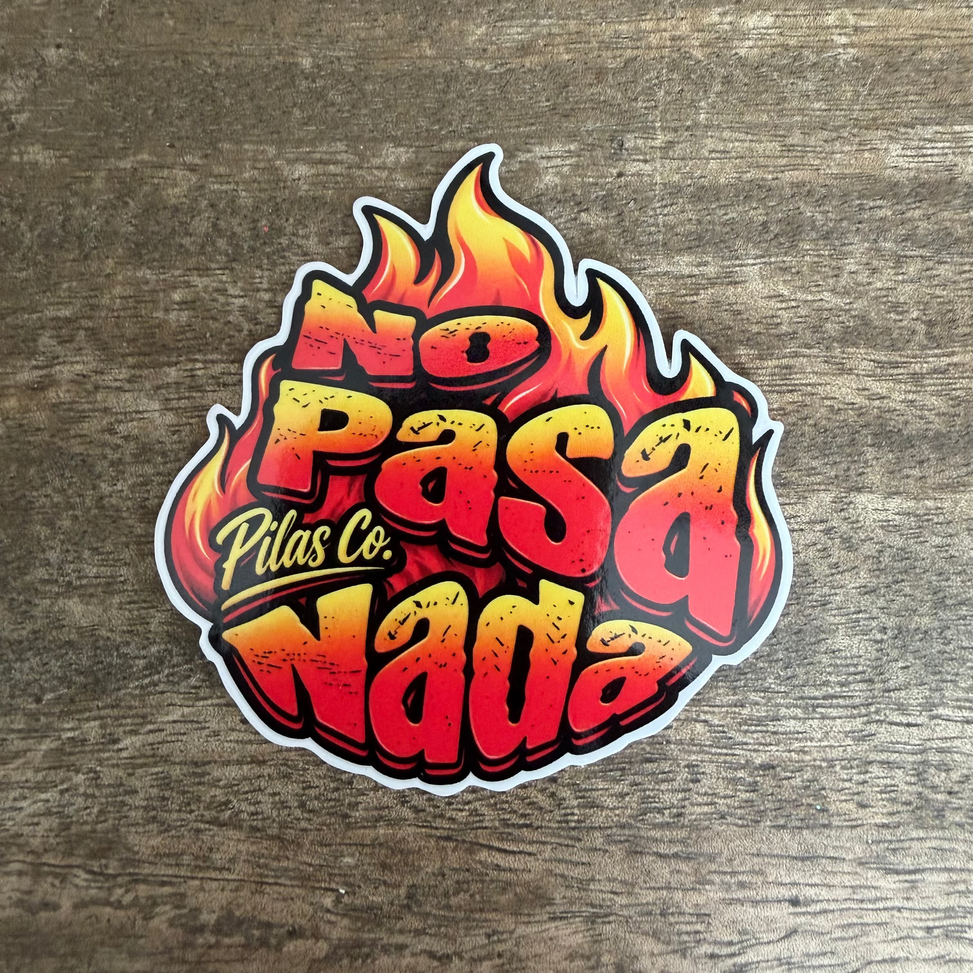 A die-cut sticker featuring the Spanish phrase 'No Pasa Nada' in bold red and yellow 3D lettering with flame graphics, placed on a brown wood-grain surface. The brand name 'Pilas Co.