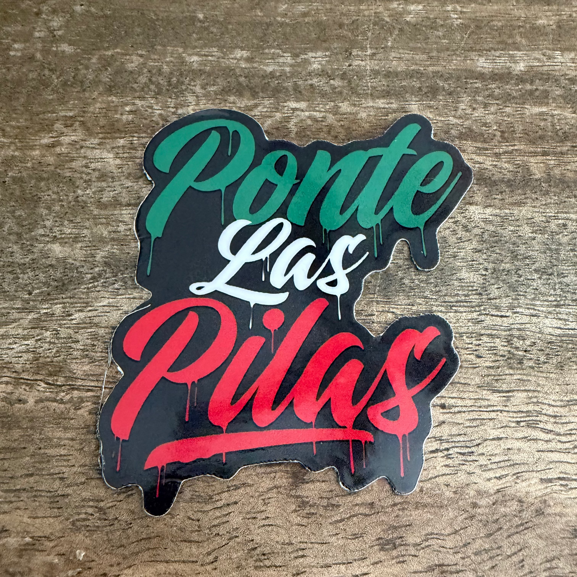A die-cut sticker with the Spanish phrase 'Ponte Las Pilas' in stylized dripping lettering, placed on a rustic wooden surface. The sticker features a black background with 'Ponte' in green, 'Las' in white, and 'Pilas' in red.