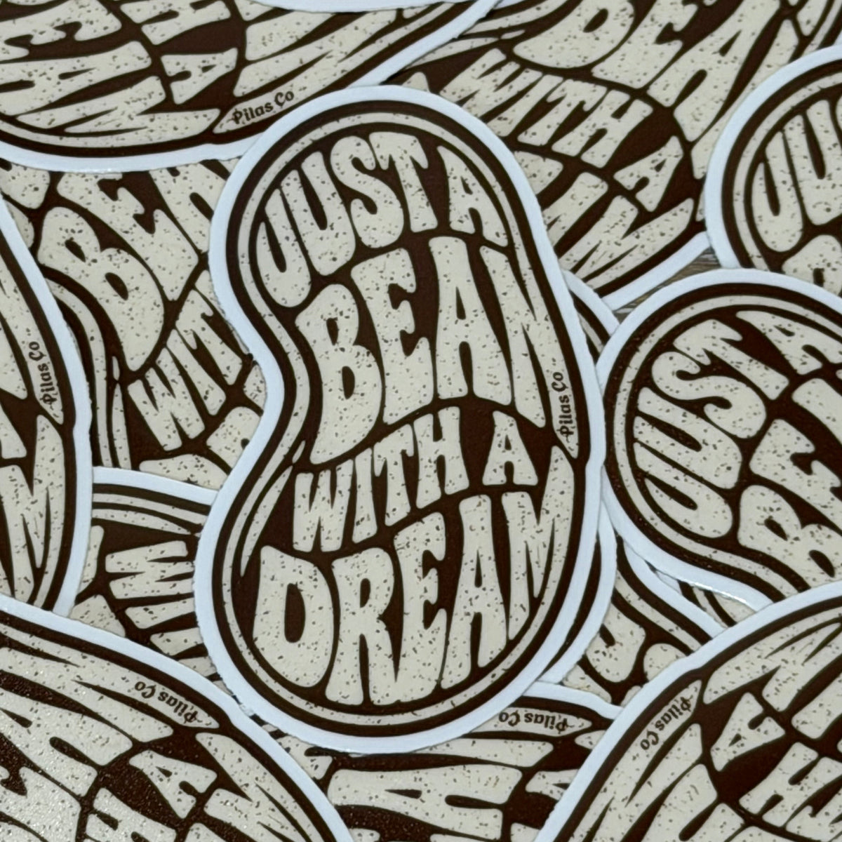 A close-up of multiple overlapping die-cut stickers featuring the phrase 'JUST A BEAN WITH A DREAM' in a textured cream font on a dark brown background, with small 'Blue Co' branding visible along the sticker borders.
