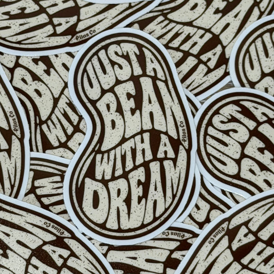 A close-up of multiple overlapping die-cut stickers featuring the phrase 'JUST A BEAN WITH A DREAM' in a textured cream font on a dark brown background, with small 'Blue Co' branding visible along the sticker borders.