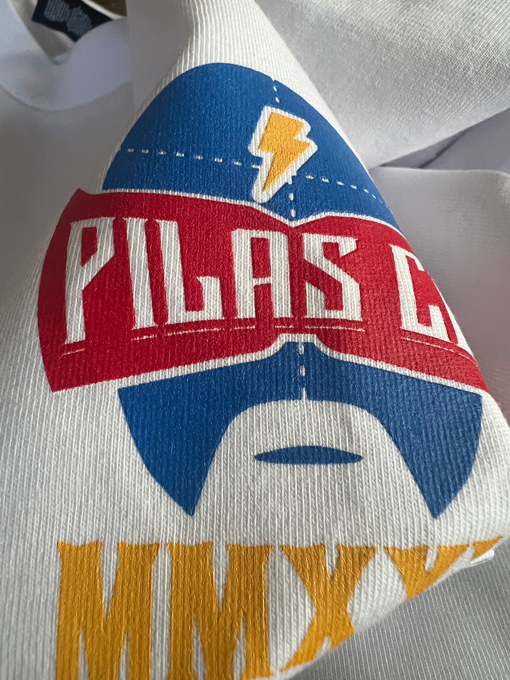Close-up detail of Pilas Clothing Co. logo shirt in white featuring blue luchador mask with lightning bolt, red banner with PILAS text, and gold/yellow accents on premium cotton fabric showcasing vibrant screen print quality